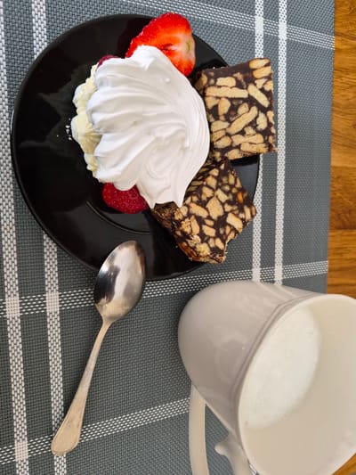 This is a top-down, close-up, high-angle photograph of a dessert and a drink on a placemat. The scene is likely indoors, possibly on a wooden table, as indicated by the visible wood grain on the right side of the image. The placemat is a dark gray fabric with a white checkered pattern.
On the left side of the placemat, on a black plate, is a serving of whipped cream with two sliced strawberries. The whipped cream is piped in a decorative swirl and appears to be very light and airy. One strawberry is sliced horizontally, showing its seeds, and the other is whole. Beneath the whipped cream, some yellowish cream or custard is visible.
To the right of the whipped cream, on the same black plate, are two pieces of a chocolate biscuit cake, also known as "hedgehog cake" or "chocolate salami." These are rectangular pieces made of broken biscuits and chocolate.
In the lower portion of the image, a silver spoon rests on the placemat. To the right of the spoon is a white ceramic pitcher filled with milk or a frothy beverage.
The lighting is bright and appears to be natural daylight. The overall composition suggests a prepared treat for consumption. There is no information in the image to indicate the specific location within Šilutė, Lithuania.