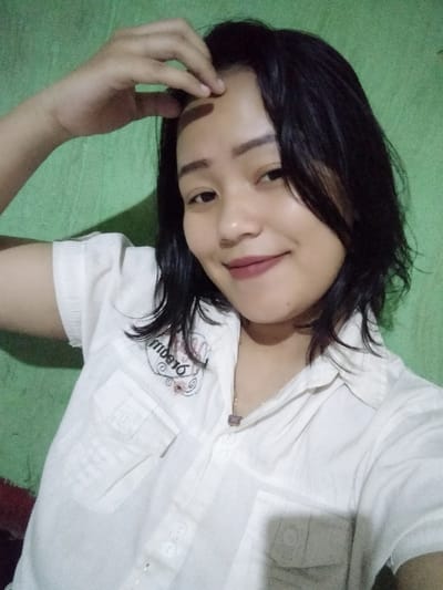 A young woman poses in a selfie, wearing a white, short-sleeved, button-down shirt. She smiles softly at the camera, her black hair falling around her shoulders. She has one hand raised to her forehead. The background is a plain, light green wall. She wears lipstick and a delicate necklace with a small pendant. The shirt has embroidered details on the left side.