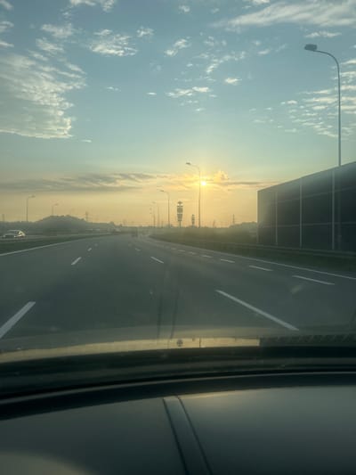 The image depicts a view from inside a vehicle driving on a multi-lane highway at sunrise or sunset. The sky is a pale blue with scattered white clouds. The sun is visible low on the horizon, casting a golden light across the scene. There are streetlights lining the highway, and a large sound barrier is present on the right side of the road. In the distance, trees and power lines are visible. The road surface is asphalt with white lane markings. The foreground shows the dashboard of the vehicle. The location is identified as a highway in or near Świętoszowice, Poland.