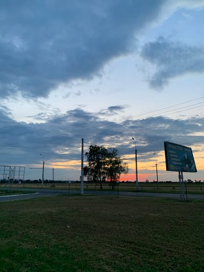 The image depicts an outdoor scene at dusk in Ternovka, Moldova. The sky is filled with dramatic, dark clouds interspersed with streaks of orange and yellow from the setting sun. The light creates a dramatic contrast with the darkening landscape.
Dominating the right side of the frame is a large billboard with text in Cyrillic. The visible text includes "ЛЮБОВЫЕ СТЕКЛО - ЗАМЕНА" which translates to "Love Glass - Replacement", and a phone number "7798-44-99, 0 6933-4-99". Below this, there are lines of smaller text that are difficult to read but appear to be services offered, such as "Тонировка" (tinting), "Химчистка" (dry cleaning), "Сигнализация" (alarms), and "Кондиционеры" (air conditioners).
In the foreground and mid-ground, there is a grassy field and a paved pathway. Several utility poles with wires stretch across the scene, indicating infrastructure in the area. A lone tree with dark foliage stands near one of the poles. In the far distance, the horizon is lined with trees and structures, suggesting a rural or semi-rural setting.
The overall mood of the image is somewhat moody and atmospheric due to the stormy sky and the fading light of dusk. The presence of the billboard suggests a commercial area, possibly along a road.