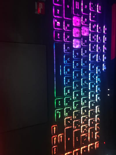 The image shows a close-up, top-down view of a laptop keyboard with RGB backlighting. The lighting shifts from red and orange on the left side of the keyboard to green, blue, purple, and pink on the right. The keys display a mix of Latin characters and what appear to be Cyrillic characters. The touchpad is visible on the left side of the keyboard, and it is not illuminated. There is a red sticker with white lettering partially visible in the upper left corner of the image, above the keyboard. The background is dark and out of focus. The image is oriented vertically with the top of the keyboard at the bottom of the frame. There is no indication of the city of Lancaster, United Kingdom, in the image.