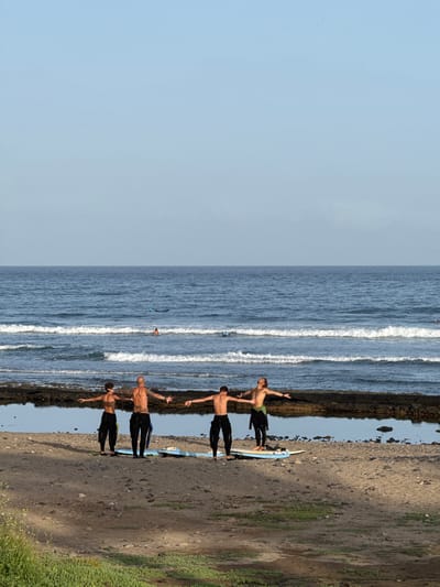 This image depicts a group of young men on a rocky beach, likely in Playa De Las Américas, Spain. The scene is bathed in what appears to be natural daylight, with clear skies and calm, blue ocean waters. The foreground is dominated by a dark, pebbly shoreline, with some sparse green vegetation growing towards the right side of the frame.
Five individuals, all appearing to be male, are standing on the shore, facing the ocean. They are dressed in black wetsuits, with some showing bare chests, suggesting they may be surfers. Each of them has their arms outstretched to the sides, their hands open, in what appears to be a group yoga or stretching pose. A light blue surfboard lies horizontally on the sand between two of the men.
In the middle ground, the ocean stretches out, with gentle waves rolling towards the shore, creating white foam lines. A few other figures can be seen in the water, one appearing to be on a surfboard, and another possibly swimming. The overall atmosphere seems peaceful and serene, with the focus on the individuals and their synchronised movements against the backdrop of the sea. There is no visible text in the image. The time of day appears to be daytime, given the lighting conditions.