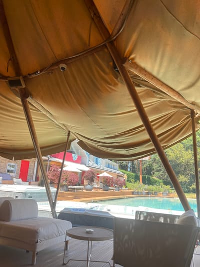 The image depicts an outdoor seating area covered by a large, light-brown tent structure with wooden poles. In the foreground, a light-colored chaise lounge sits on a wooden deck. To the right of the lounge is a small, round metal table with an ashtray. Beyond the seating area, a swimming pool is visible, with hot tubs situated near the deck. In the background, a building with red doors and white trim is partially visible, along with trees and greenery. The overall scene suggests a resort or private residence in a semi-rural or wooded setting. The location is identified as the city of Dedovo, Bulgaria.