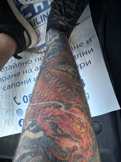The image shows a close-up view of a person's tattooed leg, which is covered in an abstract design of red, orange, and black ink. The leg is resting on a piece of paper that appears to be a business card or flyer, with blue text and logos. Some of the text is visible, including "ILIN", "ане и", "ални", and "ри". There is also a phone number starting with "08" and social media icons for Instagram and Facebook. The background is dark and out of focus, suggesting an indoor setting, possibly a vehicle or a room. The overall impression is a candid shot, focusing on the tattoo and the accompanying paper.
