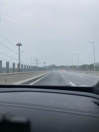 The image is a first-person perspective from inside a vehicle, looking out through a windshield covered in raindrops. It appears to be a cloudy or overcast day. The vehicle is on a multi-lane highway, with traffic visible ahead. To the left of the road, there is a row of dark, rectangular pillars. Further back, power lines and transmission towers are visible. A traffic signal with red and blue lights is mounted on a pole to the left of the road. The road surface is wet, indicating rain or recent rain. The image shows a highway scene in Luohu District, China, characterized by modern infrastructure like highways and power lines, common in urban and suburban areas.