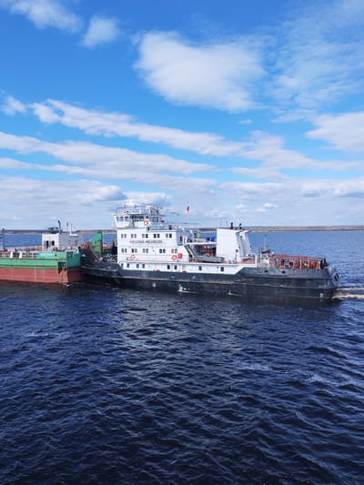 A daytime photograph captures two vessels navigating a large body of water under a partially cloudy, blue sky in Maksakovo, Russia. In the foreground, a prominent white and black ship, likely a push boat or tugboat, is actively engaged in moving a green and reddish-brown barge.

The white and black ship, named "НИКОЛАЙ МЕДВЕДЕВ" (NIKOLAI MEDVEDEV), features a multi-story white superstructure with numerous windows and a wheelhouse on top, from which a Russian flag flies. Several orange lifebuoys are mounted on its exterior. Its hull is predominantly black, with a visible wake trailing behind its stern, indicating movement.

Attached to the front or side of the "НИКОЛАЙ МЕДВЕДЕВ" is the "ВОЛЖСКАЯ-6" (VOLZHSKAYA-6), a cargo barge. This vessel has a distinctive two-toned hull: a reddish-brown lower section and a green upper section, with some white structures on its deck. The two vessels are connected, suggesting a joint operation, likely the transport of goods along a river or waterway.

The water itself is a deep blue, showing ripples and small waves, particularly around the moving vessels. In the background, a distant, flat landmass with sparse vegetation stretches across the horizon under the bright sky, which is adorned with scattered white cumulus clouds. The scene conveys a sense of active commercial navigation on a clear day.