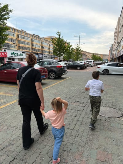 In Hunchun, China, on a cloudy day, three individuals walk away from the camera on a paved street. A woman with brown hair tied in a bun, wearing a black t-shirt and black pants, holds the hand of a young girl. The girl has blonde hair and wears a pink t-shirt and light blue pants. Behind them, a young boy in a white t-shirt and camouflage pants walks with his hands clasped behind his back. To the left of the frame, a red car is parked alongside a row of buildings. In the background, other vehicles and buildings line the street. The ground is made of interlocking concrete pavers, with a metal manhole cover visible to the right of the boy.