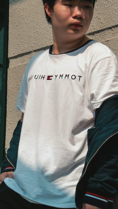 In Oreshki, Russia, a young person stands against a textured, light-colored wall. They are wearing a white t-shirt with the Tommy Hilfiger logo printed on it. The t-shirt has a dark trim around the neckline. Over the t-shirt, they are wearing a dark jacket, which is partially unzipped, revealing the white t-shirt. The jacket also has stripes on the sleeves. The lighting suggests it is daytime, with sunlight casting shadows on the wall and the person's clothing. The background is plain, with the textured wall being the dominant feature.
