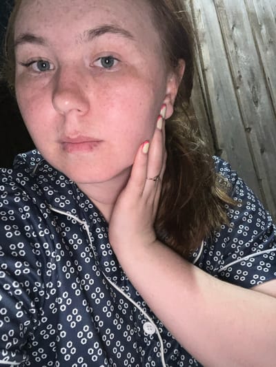 The image shows a close-up of a young woman with fair skin, freckles, and reddish-brown hair. She is wearing dark blue pajamas with a white hexagonal pattern and white piping. Her left hand is placed on her cheek, and she is wearing a silver ring on her left ring finger. Her fingernails are painted with a light beige polish and have a pink French tip. The background appears to be a wooden fence, suggesting an outdoor or rustic setting. The lighting is dim, casting shadows. There is no information available about the location in the city of Аряж, Russia.