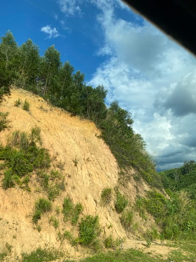 The image depicts a steep, eroded hillside in Thôn 18, Vietnam. The hillside is predominantly composed of light brown, sandy soil, with visible erosion marks and textured layers. Green vegetation, including grass and small shrubs, is growing sparsely on the slope, with denser clusters of taller grasses and bushes in some areas. At the top of the slope, a line of tall, slender trees with green foliage forms a distinct border against the sky. The sky above is a mix of bright blue with scattered white clouds and darker, more ominous storm clouds. In the background, a lush green landscape of rolling hills covered in dense trees stretches into the distance, indicating a rural or forested area. There are no people or discernible human activity in the image. The overall impression is of a natural landscape undergoing erosion, with vegetation struggling to take hold on the steep incline under a dynamic sky.