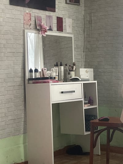 The media file shows a close-up, rotated view of a vanity setup, likely in a home setting. The scene is dominated by a white makeup organizer with multiple compartments, some of which are filled with various cosmetic products like brushes and bottles. To the left of the organizer, a large mirror reflects the contents of the vanity and the surrounding wall, which is clad in light grey brick-like tiles. A few decorative items, including a pink orchid and some patterned paper squares, are placed on the wall near the mirror. In the background, a wooden chair with a dark seat is visible, positioned next to a table. The overall impression is of a personal grooming or makeup station. The image does not depict any people. No specific time of day, weather, or strong emotional cues are apparent, other than a sense of organized personal space. There is no visible text in the media file. The provided location context is Линёвка, Russia, which might indicate the geographical setting of this scene.