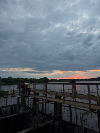 The image depicts a group of people standing on a metal structure overlooking a body of water, likely a river, at dusk. The sun is setting on the horizon, casting a warm glow of orange and pink across the sky, which is otherwise filled with dramatic, dark gray clouds. The setting appears to be natural, with dense green trees lining the opposite bank of the water. The metal structure the people are on has a somewhat industrial or utilitarian appearance, with visible beams and supports. Some of the people are looking out at the sunset, suggesting they are enjoying the view. The overall mood of the image is serene and contemplative, enhanced by the natural beauty of the sunset and the quiet atmosphere.