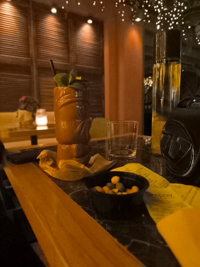 The image presents a close-up, dimly lit scene on a table within an indoor bar or restaurant in Palaio Faliro, Greece, likely during the evening. The central subject is a distinct brown and orange tiki mug cocktail, garnished with fresh mint leaves and a small yellow flower, with a dark straw protruding. It sits on a crumpled yellow napkin on a dark, marble-patterned table.

To the right of the cocktail, an empty, clear glass tumbler is placed. Further right, a tall, transparent bottle filled with a yellow liquid stands next to a black, compact object resembling a speaker or a part of a camera. In the foreground, a small black bowl contains an assortment of green and yellow/brown spherical snacks. Several crumpled yellow paper receipts are visible on the table, one displaying the partial text "epay." A warm, wooden surface extends into the frame from the lower-left.

The background is softly blurred, showing horizontal wooden slat-like walls, an orange-hued pillar, and numerous small, sparkling lights that create a bokeh effect, suggesting fairy lights or decorative lighting overhead. A faint, glowing light, possibly from a candle or small lamp, is visible on another table in the distance, alongside a blurred figure. The scene evokes a relaxed, social atmosphere.