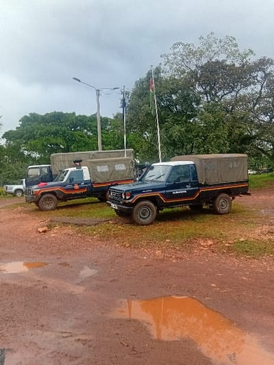 The image depicts an outdoor scene in Shirungu-Mugai ward, Kenya, characterized by an overcast sky and wet, reddish-brown dirt ground with multiple standing water puddles. In the foreground and midground, two dark blue police utility vehicles, identified as Toyota Land Cruiser-type pickups, are parked. The vehicle in the foreground has a canvas cover over its bed and displays "POLICE" text along with red, yellow, and blue stripes. The second vehicle, positioned behind and slightly to the left, also has a white cab, a dark canvas-covered bed, "POLICE" text, and a red emergency light visible on its roof. A third, lighter-colored utility vehicle is partially visible in the far background. A tall flag pole bearing the Kenyan national flag is visible in the midground, with a utility pole equipped with light fixtures situated to its left. Lush green trees and foliage form a dense backdrop. No individuals are visible in the scene.