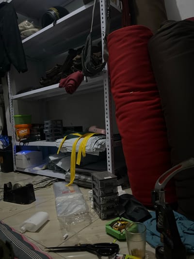 The image displays a cluttered storage area, possibly a garage or workshop, with various items stacked on metal shelving units. In the foreground, large cylindrical objects, one red and one dark-colored, rest on the top shelf. Below them, a variety of items are visible, including what appears to be a black motorcycle helmet with yellow stripes, some rolled fabric, and what looks like a collection of gloves.

Further down the shelving, there are several smaller containers, some green and orange, one labeled "LOVY" with text indicating "Assorted Cookies." A white electronic device with a blue light is also present. Towards the right side of the image, a glass with a light-colored liquid sits in a green ashtray filled with cigarette butts. Nearby, a pair of large black scissors and a white cylindrical object are placed on a surface. A large clear plastic-wrapped item is also visible.

The lighting suggests it might be indoors with artificial light, and the overall scene conveys a sense of disarray and storage of miscellaneous goods. There are no people visible in the image. The environment appears to be a domestic or commercial storage space. Some text is visible on the "LOVY" packaging.
