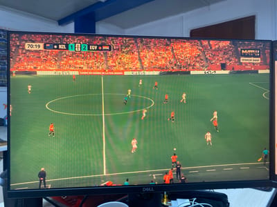 The image displays a close-up view of a computer monitor showing a soccer match being broadcast. The screen shows a scoreboard with the time "70:19", the teams "NZL" (New Zealand) and "EGY" (Egypt), and the score "1-2" in favor of Egypt. The stadium is filled with spectators wearing predominantly orange clothing. Advertising banners for "Kia EV5" and "FIFA" are visible. The game appears to be taking place on a green field with white markings. Several players in orange and white uniforms are on the field, along with referees. A logo for "МАТЧ!" with the text "ПРЯМОЙ ЭФИР" (Live Broadcast) is present on the right side of the screen, indicating the broadcast channel. The monitor is situated on a desk with some cables and what appears to be a portion of another monitor to the right. The background suggests an indoor environment with a blue ceiling structure and a wall. While the image shows a digital representation of a soccer match, there is no direct indication of the city of Yukhta, Russia, within the visual content of the screen or the immediate surroundings captured by the camera.