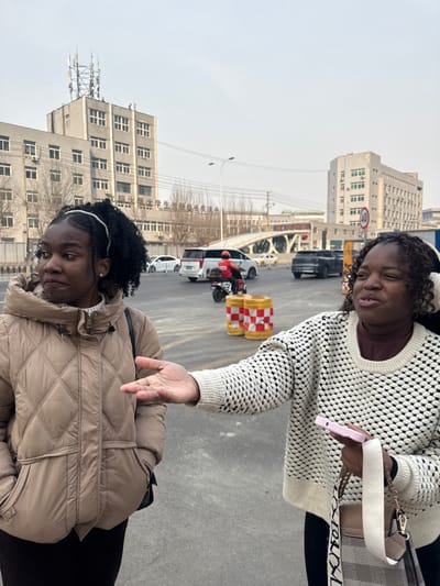 The image captures an outdoor urban street scene in Dadong District, Shenyang, China, under an overcast sky. Two females with dark skin are prominent in the foreground. The female on the left wears a light-tan quilted hooded winter jacket over a dark top, with dark curly hair secured by a white beaded headband. She has her left hand in her pocket and is looking slightly to her left. The female on the right wears a cream-colored textured knit sweater over a brown high-neck top, with a white accessory, possibly earmuffs, on her head. She holds a pink smartphone in her right hand and carries a beige and white patterned shoulder bag with "XOXO" text on the strap. Her left arm is extended forward with an open palm, gesturing as she speaks.

In the mid-ground, a paved road exhibits vehicular traffic, including a white SUV and a dark SUV. A person wearing a red helmet and red jacket is riding a red electric scooter/motorcycle, moving from left to right. Two red and white striped cylindrical construction barriers are visible on the road. A "40" speed limit sign is mounted on a pole to the right. The background features multiple multi-story grey concrete buildings; the prominent building on the far left has communication antennas on its roof and is identified by Chinese characters as "Yangtai Vocational and Technical College Affiliated Secondary Vocational School." An arched bridge is also visible in the background. Sparse, bare trees are scattered throughout the scene.