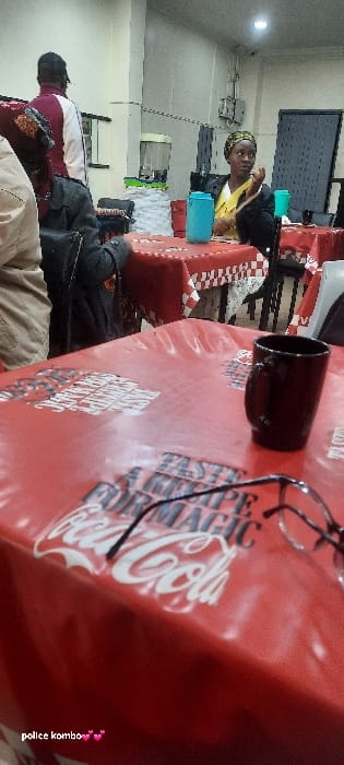An indoor scene depicts a dining or communal area within a commercial establishment in Homa Bay, Kenya. In the foreground, a red tablecloth emblazoned with white Coca-Cola branding and the text "TASTE A RECIPE FOR MAGIC" covers a table. A black ceramic mug and a pair of dark-framed eyeglasses rest on this tablecloth. In the mid-ground, a dark-skinned woman with a yellow top, dark jacket, and patterned headscarf is seated at a table, looking left with her right hand near her chin. Two light blue insulated water bottles are on her table. To her left, a person stands with their back to the camera, wearing a maroon and white top and a dark cap. Another person is partially visible seated with their back to the camera, wearing a dark jacket. In the background, a transparent beverage dispenser containing ice and liquid stands on a counter, flanked by stacks of cups. The walls are light-colored, and the area is illuminated by overhead artificial lights. Electrical conduits are visible on a wall in the background, next to a dark panel or door. Other tables with red and white checkered tablecloths and dark chairs are visible further back.