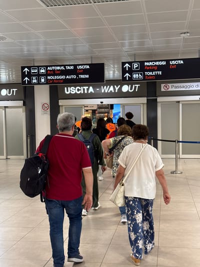 The image shows a queue of people standing in what appears to be an airport or train station. There are multiple signs directing people to different services: buses and taxis, car rentals and trains, parking, toilets, and the exit. The signs also include the Italian words for these services: 'Autobus, Taxi,' 'Noleggio Auto, Treni,' 'Parcheggi,' 'Toilette,' and 'Uscita' (Exit). Some people are waiting near a barrier with a sign saying "Passaggio di." The location is likely in Cinisi, Italy, as the signs are in Italian. The photo captures travelers following directions at an airport or transportation hub.