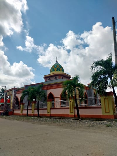 The image shows a mosque in Labissa, Indonesia. The mosque has a prominent dome with a green and yellow checkered pattern, surrounded by a pale pink building with orange trim. There are palm trees in front of the mosque, and a metal fence separates it from the street. The sky is blue with scattered white clouds. A utility pole is visible on the right side of the image. The street in front of the mosque is paved with asphalt and appears to be dusty. A sign on the left side of the mosque reads "MASJID JAMI NURUL YAQIN".
