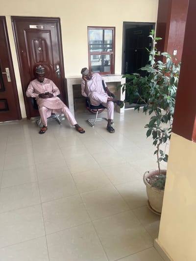 Two men dressed in similar light pink traditional attire are seated on metal chairs in an outdoor courtyard in Jama A, Nigeria. Both men wear traditional caps and sandals. One man is holding a cell phone. The courtyard has beige tiled flooring and walls. A potted ficus tree stands to the side. A dark brown door is visible on one wall, and a black door and a window are seen above it.