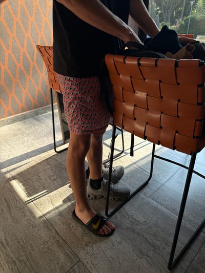 A man wearing a black t-shirt and red patterned shorts is sitting in an orange woven chair. He is wearing grey sneakers on one foot and a black Nike sandal on the other. The chair is on a tiled floor in an interior space. Another area of the floor is covered in orange and grey patterned flooring. The location is Santa Ana Yancuitlalpan (Chilhuacán), Mexico.