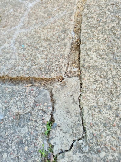 A close-up, overhead shot captures the textured surface of cracked concrete slabs. The concrete is a mix of gray aggregate and smaller pebbles of various colors, including pink, white, and dark gray. A prominent crack runs diagonally from the upper right to the lower left, bisecting the image. Another crack forms a perpendicular line across the center of the image, intersecting the first. Small patches of green grass are emerging from the cracks in the lower left and right portions of the frame. The scene is likely a section of pavement or a sidewalk. The location is an unspecified outdoor area in Švenčionys, Lithuania.