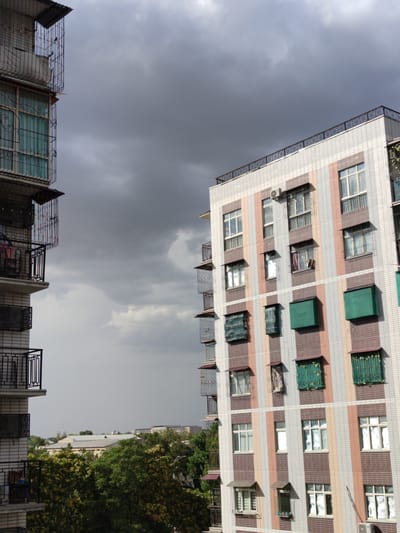 This image shows an exterior view of two apartment buildings in Minglola mahalla, Uzbekistan, under a cloudy sky. The building on the right is a modern, multi-story structure with a facade of pink, white, and brown tiles arranged in vertical stripes. It features numerous windows, many with green awnings or metal coverings. Balconies with black railings are visible, some with laundry hanging. An air conditioning unit is mounted on the building's exterior.

The building on the left appears older and is characterized by its light-colored brickwork and extensive use of decorative metalwork on its balconies and window guards. Laundry is also visible hanging from some of these balconies.

In the background, lower buildings and lush green trees are visible, suggesting a residential area within the city. The sky is overcast, indicating potentially poor weather. No people are visible in the image.