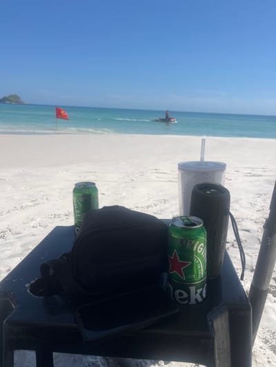 A beach scene with a table holding a clear cup with a straw, a black speaker, two green Heineken cans, a black bag, and a smartphone. In the background, the ocean meets a sandy beach under a bright sky. A red flag is visible on the beach near the water's edge. Further out in the water, a red and grey watercraft can be seen. The photo has been taken at a tilted angle.