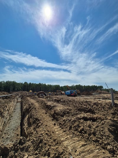 The image shows an outdoor construction site in Taezhnoe, Russia. The sun is high in the sky, creating a bright glare. The sky is a vibrant blue with wispy white clouds. The foreground of the image is dominated by a large trench filled with gravel. The ground is composed of dark brown soil, with visible tire tracks from heavy machinery. In the middle ground, there are various construction vehicles, including a blue and red tracked vehicle with the text "2450 CH-24" visible, an excavator, and other machinery. Large pipes are also visible, including a large curved pipe and several smaller ones. A line of trees forms the background of the scene, suggesting the site is near a forest. A tall, grey pole stands on the right side of the image. The overall impression is one of active construction and land development.
