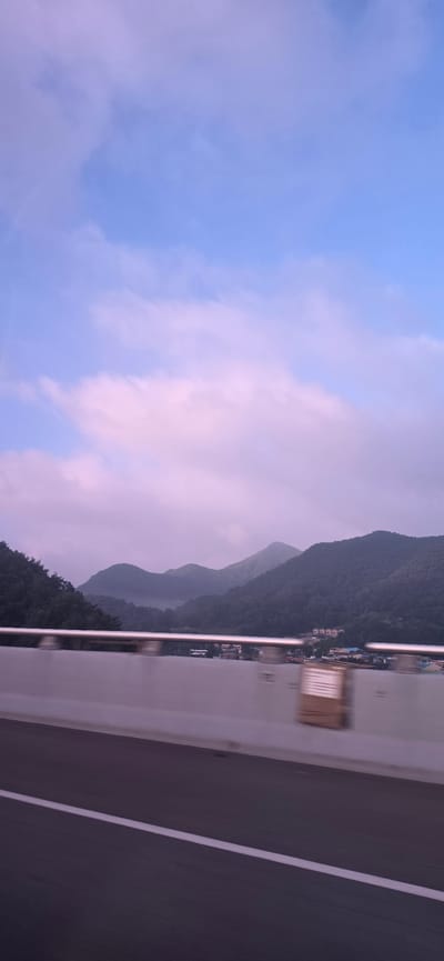The image depicts a scenic view of a mountainous landscape with a clear sky and scattered clouds, likely taken from a moving vehicle on a road. The foreground features a road with a white line marking the lane, suggesting it's a highway or a major road. A guardrail runs horizontally across the middle of the frame, separating the road from the view beyond. Behind the guardrail, a town or village is nestled at the base of lush, green mountains. The mountains are covered in trees and have rolling contours, with a hint of mist or fog in the valleys. The sky is a soft blue with streaks of pink and white clouds, indicating either sunrise or sunset. The overall impression is one of natural beauty and tranquility, possibly captured in Hoein-myeon, South Korea, given its mountainous terrain and typical rural architecture visible in the distance.