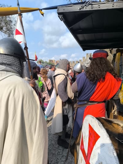 The image depicts a scene at the medieval festival in Óbidos, Portugal. In the foreground, a person in a white cloak and chainmail hood is facing away from the camera. To their right, a man with a beard wears a chainmail hood and a long, cream-colored tunic. He is holding a wooden staff. Further to the right, a person in a blue and red tunic with a red and blue turban stands with their back to the camera. In the background, there are people dressed in medieval attire, and a stone wall, characteristic of Óbidos Castle, is visible. A flag with a red cross on a white background is prominent in the upper left portion of the image. The ground appears to be paved with cobblestones. The sky is blue with scattered clouds.