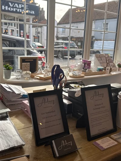 The image presents an interior view from a shop named "Alchemy At Home Inspired Living," located in Hornsea, United Kingdom, looking out onto a street scene. In the foreground, a wooden counter features two black-framed signs. The left sign details a "Get Rewarded" loyalty program, inviting customers to pick up a loyalty card, receive a stamp for every £20 spent, and redeem a full card for £20 off. Below this sign is a stand holding "Alchemy At Home Inspired Living" loyalty cards. The right sign outlines the "Refunds & Exchange Policy." To the left, a stack of papers and documents is visible. A black cash register or point-of-sale system is situated between the two framed signs. Small business cards or flyers are placed on the right side of the counter.
The midground is dominated by a white-framed window with multiple panes, acting as a display ledge. Various homeware items are arranged here, including a small green potted plant, assorted clear glass and crystal bowls, glasses, a decanter, and a rectangular light-colored tray. A prominent dark blue abstract sculptural object stands near the center. Further to the right, a pink-tinted large goblet, an air freshener or diffuser with reeds, and other small decorative items like candles are displayed.
Through the window, the exterior streetscape of Hornsea is visible under bright, overcast daylight. On the left, a black SUV is partially parked, and a shop sign reading "VAPE Hornsea" is clearly identifiable on a building across the street. Further down, traditional brick buildings and other vehicles are present. On the right, a grey SUV is parked, followed by a white delivery van. Several light-colored buildings line the street. Reflections of interior objects are visible on the windowpanes. No individuals are depicted within the scene.