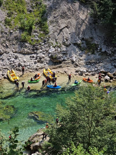 This aerial, high-angle drone shot captures a scenic view of the Enns River near Landl, Austria, during a sunny day. The river, with its crystal-clear, emerald green water, flows through a rocky, mountainous terrain. Along the riverbanks, lush green foliage and trees contrast with the grey, rocky slopes.
Several individuals are engaged in water sports. In the foreground, a large, vibrant green tree partially obstructs the view of the water. On the rocky bank, a person in black clothing stands near a large rock formation. A group of people in wetsuits and life jackets are preparing for or have just completed water activities.
There are multiple inflatable rafts and kayaks scattered around. Two yellow inflatable rafts are partially submerged in the shallow water along the bank, each carrying several people. A bright orange inflatable kayak with one occupant is positioned between the yellow rafts. A bright blue inflatable kayak with one person is also in the water. A smaller, lime green inflatable kayak with one person is near the orange kayak.
Further upstream, more people in wetsuits and helmets are visible, some standing on the bank and others in the water. The rocky bank is covered with loose stones and larger boulders. The overall scene depicts a group enjoying outdoor recreational activities on a river in a mountainous region.