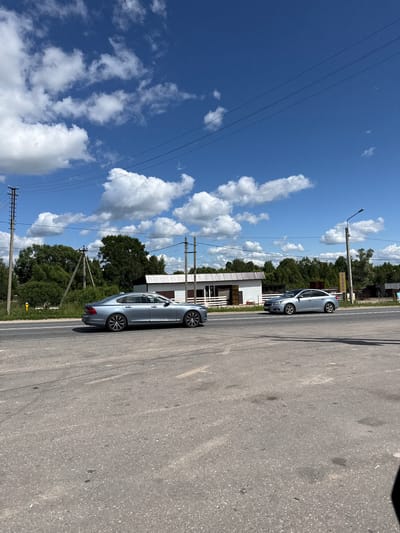 In Staraya, Russia, on a sunny day with scattered white clouds against a bright blue sky, a silver sedan is seen driving on an asphalt road. The car is positioned in the left lane, facing right. In the background, there is a white building with a dark roof, several green trees, and utility poles with power lines. Another silver car is parked on the right side of the road, facing left. The road appears to be a two-lane street, with white lane markings visible. A lamppost stands on the right side of the road, and a yellow marker is visible on the left side. The overall scene depicts a rural or suburban setting with a focus on road traffic and infrastructure.