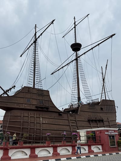 The image depicts a large, dark brown wooden replica of a multi-masted sailing ship, identified as the Maritime Museum (Kompleks Muzium Samudera) located in Malacca City, Malaysia. The ship, a prominent structure, features a planked hull and three visible masts equipped with intricate rigging and rope ladders; no sails are deployed. The scene is observed under an overcast sky.

Approximately ten individuals are positioned on the ship's upper deck on the right side, with two additional individuals on a lower deck, appearing to move and observe their surroundings.

In the immediate foreground, a low, red and white painted concrete barrier with decorative pillars extends horizontally. Several potted plants displaying purple flowers are arranged along this barrier. To the far right, a small red brick building is visible, bearing a sign in white lettering that reads "KOMPLEKS MUZIUM SAMUDERA." On the paved ground in front of the barrier, two adult males are walking from left to right: one in a patterned shirt and dark trousers, and another in a light-colored shirt and blue jeans, looking down at a mobile device. A black and white striped curb delineates the pavement.