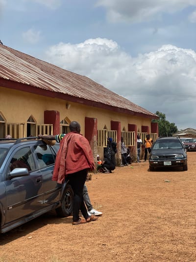 An outdoor, eye-level, medium shot captures a group of people standing and sitting outside a long, single-story building with a corrugated metal roof. The building's exterior walls are painted a pale yellow with dark red columns at intervals. Windows with barred openings are visible along the building's facade. The ground is covered in reddish-brown dirt.

In the foreground, a dark grey car is parked on the left side of the frame, facing right. A man in a red jacket and black pants stands to the right of the car, leaning on its roof. His back is to the camera.

To the right of the car, a black sedan is parked facing the camera. Its license plate reads "PKN 631 KM". Other cars are visible further in the background.

A group of people are gathered near the building, some standing, some sitting on the ground. They appear to be engaged in conversation or waiting. The sky above is partly cloudy. The location is Kassa, Nigeria.