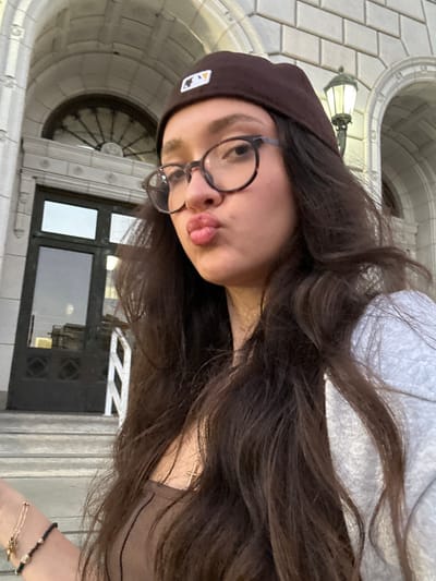 A young female is depicted in a selfie, posing with pursed lips, wearing a dark brown beanie with a white MLB logo and round tortoiseshell-patterned glasses. She has long, dark brown hair and is dressed in a brown top and a light grey hooded sweatshirt, accessorized with a gold cross pendant necklace and multiple bracelets on her left wrist. The background features the exterior of the Glendale City Hall, located at 613 E Broadway, Glendale, CA 91206. Visible architectural details include a light-colored stone facade with a large arched entrance, dark multi-paneled double doors, a semi-circular fanlight window with radial spokes above the doors, and a traditional-style street lamp mounted on the building's right side. White stone balustrades indicate steps leading up to the entrance. The scene is illuminated by natural light.