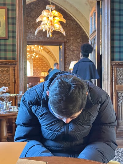 The image depicts an ornate interior within the Abbaye des Vaux de Cernay, located in Cernay-la-Ville, France.
In the immediate foreground, a young individual with short, dark hair is seated at a polished dark wooden table, their head bowed downwards, looking intently at the table surface. This person wears a black puffed winter jacket featuring a circular logo emblem on the left chest and a blue neck gaiter or scarf. To the left of this individual, on a separate round wooden table, a two-tiered silver-colored serving stand holds small, light-colored food items such as pastries or snacks. Surrounding the stand are several small white dishes, jars, and a glass bottle. A hand, likely belonging to another unseen individual, is visible near these items.
The background reveals the historic and luxurious architecture of the establishment. Walls are adorned with dark, intricately carved wood paneling and sections of dark green tartan fabric. Portions of the wall feature patterned wallpaper with stylized motifs in muted gold and brown tones. A large, ornate chandelier, composed of multiple tiers of illuminated, possibly crystal, leaf-like elements, hangs from the high ceiling, providing warm lighting. To the right of the background, a person with dark hair styled in a bun is visible from behind, wearing a dark cape-like garment, standing near a wooden doorway. Through a large archway or a mirror framed as an archway in the center background, an adjacent room with similar decor, including another chandelier and a window, is visible. Within this reflected or connected space, a man in a light-colored top is seen standing, facing away from the camera, alongside other indistinct figures. A leopard-print floor covering is partially visible on the right side of the foreground.