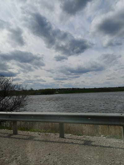 This media file captures a wide landscape scene featuring a body of water, land, and an overcast sky, likely taken from a roadside perspective in Sofiivka, Ukraine.
In the foreground, a gray metal guardrail stretches horizontally across the lower part of the frame, indicating the edge of a road or viewing area. Below the guardrail, there is a visible gravel or dirt road shoulder. Behind the guardrail, some dry, light-colored reeds or tall grasses can be seen near the water's edge. To the left, a portion of a tree with bare, dark branches extends into the frame.
The midground is dominated by a large body of water, possibly a lake or wide river. The water's surface is textured with numerous ripples and small waves, suggesting windy conditions.
In the background, on the opposite bank of the water, a relatively flat expanse of land is visible. This land features patches of green grass and scattered trees, indicating a natural or agricultural landscape. Several small, distant buildings, likely houses, are dotted along the far shore, suggesting a rural settlement or village.
The sky is heavily overcast with dense, grey clouds that fill the upper portion of the image. While the clouds are predominantly dark grey, some lighter grey patches suggest varying thickness or breaks in the cloud cover. There is no direct sunlight visible, indicating a cloudy daytime.
No specific human or animal activity is depicted. There is no legible text visible in the image. The overall impression is one of a quiet, rural landscape on an overcast day.