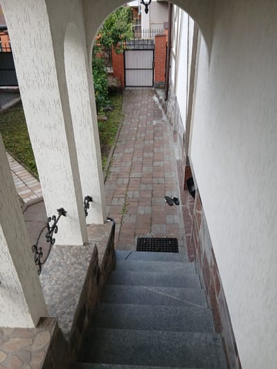The image shows a view from an arched entryway looking down a paved path towards a gate. The entryway has thick, textured white columns. To the left of the path is a patch of green grass, and to the right is the textured wall of a building. The path is made of interlocking pavers and is lined with green grass. Further down the path, there is a metal gate with vertical bars and a solid metal panel, set between brick pillars. A tree with green leaves is visible behind the gate. There is a pair of dark-colored flip-flops on the paved path near a black doormat. The scene is set in Vita-Poshtova, Ukraine. The perspective suggests the photograph was taken from a slightly elevated position, possibly a porch or balcony.