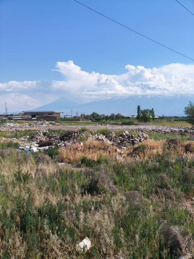 This image depicts an outdoor scene in Nor Kharberd, Armenia, on a bright, sunny day. The foreground features an unkempt field dominated by a mixture of dry, light-brown grasses and green weeds. Scattered litter, including white and blue plastic bags, is visible among the vegetation. In the midground, a significant accumulation of refuse and debris is present, consisting of various rocks, twisted metal, and discarded materials. Behind this debris field, a low-slung, light-colored building with a dark roofline and some support structures is partially visible. Further back, several power line towers are present. The background is dominated by two prominent snow-capped mountains, likely Mount Ararat, partially obscured by large white cumulus clouds against a clear blue sky. A single black power line traverses the upper right portion of the frame diagonally. No individuals are present in the scene.