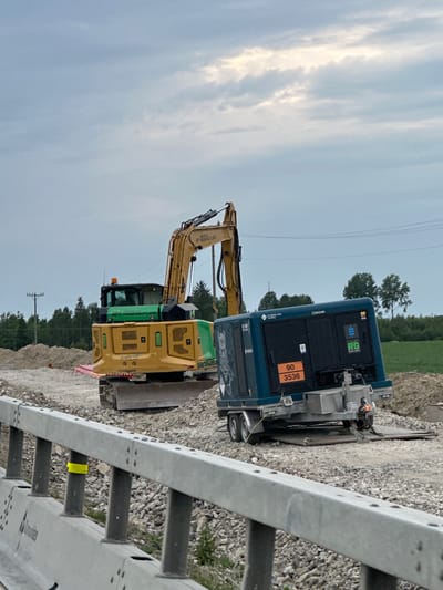 The image shows a construction site in Slitu, Norway, with two pieces of heavy machinery: a yellow excavator and a blue generator unit. The excavator is positioned in front of the generator, with its boom arm extended upwards and to the left. The generator is a large, blue, trailer-mounted unit with its doors closed.

The scene is set outdoors, with a gravelly ground surface and piles of dirt. In the background, there are trees and a green field, suggesting a rural or semi-rural location. Overhead, a cloudy sky dominates the upper portion of the image. The lighting suggests it is daytime, likely late afternoon or early evening due to the diffused light and cloud cover.

Notable details include a hazard sticker on the generator with the numbers "90 3536" and what appears to be a company logo with "RG". There are also power lines visible in the background, indicating proximity to electrical infrastructure. A metal railing with regularly spaced holes runs along the right side of the image, possibly part of a roadside barrier or construction scaffolding.

There are no people visible in the image. The overall impression is one of a quiet, possibly paused, construction operation under an overcast sky. The image is oriented vertically.