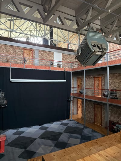 The image shows the interior of a large, industrial-style space, likely a studio or performance venue, located in Shabdan, Kyrgyzstan. The space features exposed brick walls, metal beams and supports forming multi-level walkways and balconies, and a high ceiling with skylights. A large, green theater spotlight is suspended from the ceiling. In the foreground, a section of the floor is covered with a checkered pattern of dark and light gray tiles. A black curtain hangs on the left side of the frame, and wooden bleachers are visible in the lower right corner. Several wooden doors with metal hinges and latches are set into the brick walls on the various levels. Small spotlights are mounted on railings along the balconies. The overall impression is of a functional, somewhat raw, and adaptable space. There are no people visible in the image.