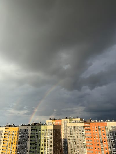 The image captures a view of a city in Анкудиновка, Russia, under a cloudy sky with a faint rainbow stretching across the scene. High-rise apartment buildings stand to the right, featuring varied color palettes including orange, gray, brown, green, and yellow. The sky is predominantly gray with some lighter patches where the rainbow is visible.