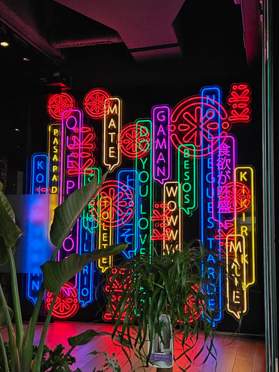 This media file presents a vibrant, multi-colored neon sign display illuminating a dark interior, likely a restaurant, bar, or entertainment venue in Santa Cruz De Tenerife, Spain. The signs feature a lively mix of text in various languages and stylized circular patterns resembling flowers or wheels.
The predominant colors of the neon lights are red, blue, green, yellow, pink, and purple, arranged in a dynamic, overlapping composition. Visible English text includes "Kiki," "GUESTARE," "BESOS," "WOWWW," "GAMAN," "DO YOU LOVE:", "MATE," "PASAPAD," "KO," and "RIO." A purple neon sign displays Japanese characters "食欲が旺盛," meaning "good appetite" or "hearty appetite."
Lush green foliage from several plants partially frames and obscures parts of the neon art, adding a natural contrast to the artificial glow. A plant pot is visible on the right, bearing the label "BEAUCARNEA" with smaller text "Jubas Palm." The scene is brightly lit by the signs themselves, casting colorful hues on the dark background and creating an energetic, modern, and inviting atmosphere. The image captures the aesthetic of a trendy, urban establishment. No people or specific activities are depicted, focusing solely on the decorative lighting and botanical elements.