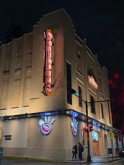 The Olympia Theater's exterior at night showcases its illuminated signs. Two neon signs with the name "Olympia" adorn the building's facade. Above, another sign displays the theater's name. Street-level, decorative neon spirals are visible. A traffic light displays green. A pedestrian crossing sign is visible near the top right corner. Sidewalk pedestrians walk along the building. The building features recessed panels and windows, creating a textured appearance.
