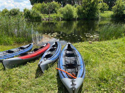 Four inflatable kayaks are positioned on a grassy bank next to a calm body of water. From left to right, the kayaks are colored blue and grey, red and grey, and two are blue and grey. The red kayak has "TRITON" written on its side. An orange pole with a rope is inside the rightmost kayak. The water is still and reflects the surrounding greenery and a cloudy sky. Tall grasses and lush trees line the opposite bank of the water, forming a dense natural boundary. The overall setting is a serene, outdoor location, possibly a river or lake, in a natural environment. This scene appears to be located in Shchuchye, Russia, given the context provided. There are no people visible in the image.