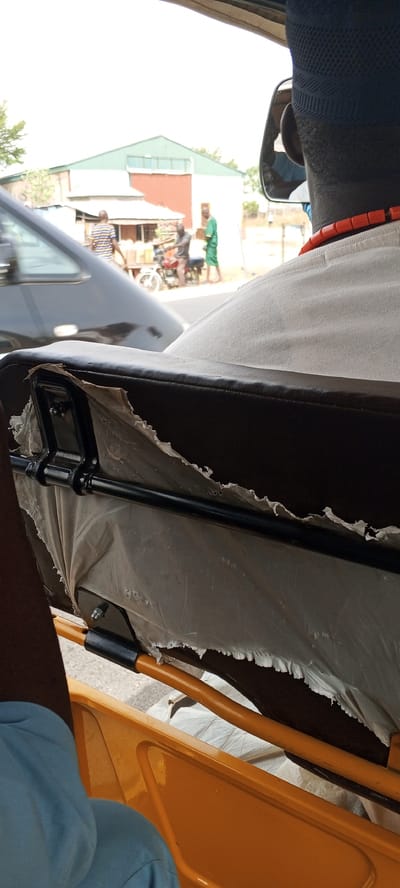 The image, taken from inside a vehicle, shows the back of a person wearing a black cap and a red beaded necklace, presumably the driver or passenger. The torn seat of the vehicle is in the foreground. Through the vehicle's windows, a street scene unfolds. There is a black car on the left and, further in the background, a motorcycle with two people on it and another person standing nearby. A building with a green and red roof is visible in the background. Based on the metadata, this scene is located in Uke, Nigeria.