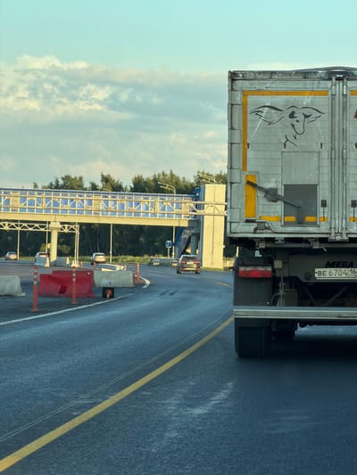 The image depicts a view from a vehicle traveling on a wet, asphalt road, likely a highway or a toll road, with a clear yellow line marking the lane. In the foreground, the rear of a large, silver truck with a stylized image of a bull or cow on its side is visible. The truck's license plate reads "BE 6704 164 RUS". The truck also has "MEGA" printed on its rear. The road is lined with temporary construction barriers, including red and white concrete barriers and orange traffic cones, suggesting ongoing work or a diversion. In the mid-ground, several passenger cars are driving in the same direction. A large pedestrian bridge with blue paneling spans across the road in the distance. Beyond the bridge, a dense line of green trees forms the horizon. The sky is a pale blue with scattered, fluffy white clouds, indicative of fair weather, possibly early morning or late afternoon given the soft lighting. The overall scene is consistent with a transport hub or a section of road undergoing maintenance within or near the city of Egor'evo, Russia.